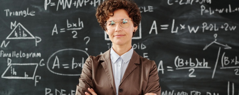 A teacher in a brown suit in front of a black board covered in equations, showing her motivation to teach.