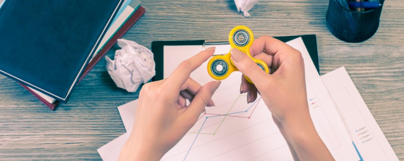 A pair of hands with a yellow fidget spinner, representing a student with ADHD using one in the classroom. Books and papers are stacked around.